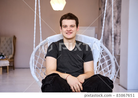 Smiling 23 years old man in hanging chair indoors. Smiling 23 years old man in hanging chair indoors. 83867651