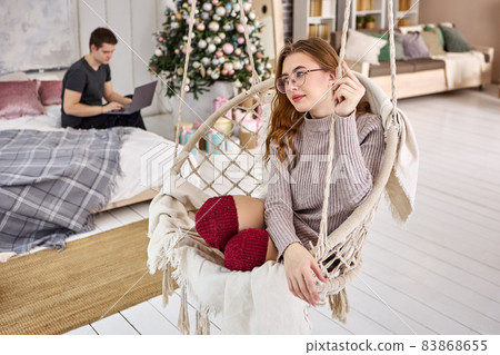 Hanging chair in New Year interior, young woman swinging on swing, man on bed with laptop. Hanging chair in New Year interior, young woman swinging on swing, man on bed with laptop. 83868655