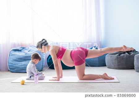 A pregnant woman in sportswear does gymnastics while her son plays in the mobile. Girl on yoga with her child. Fitness mom and baby. A pregnant woman in sportswear does gymnastics while her son plays in the mobile. Girl on yoga with her child. Fitness mom and baby. 83869800