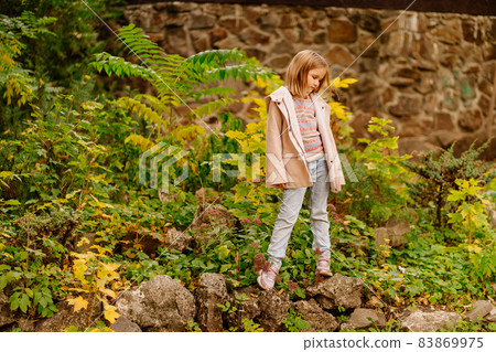 a cute little girl in a coat and jeans in an autumn park with yellowing foliage. a cute little girl in a coat and jeans in an autumn park with yellowing foliage. 83869975