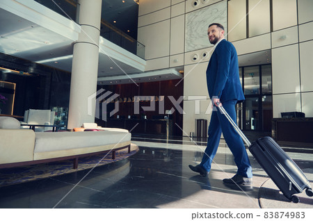 Handsome man, successful business developer smiling, looking to the side, walking down the lobby, heading with luggage to the lounge area inside a modern luxury building during at trip Handsome man, successful business developer smiling, looking to the side, walking down the lobby, heading with luggage to the lounge area inside a modern luxury building during at trip 83874983