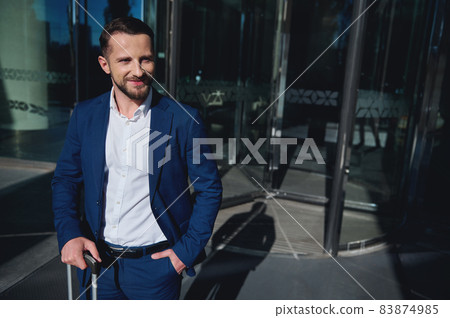 Self-confident businessman developer entrepreneur in business suit looks aside posing with a luggage and his hand in pocket on the background of corporate modern high-rise building glass entrance Self-confident businessman developer entrepreneur in business suit looks aside posing with a luggage and his hand in pocket on the background of corporate modern high-rise building glass entrance 83874985