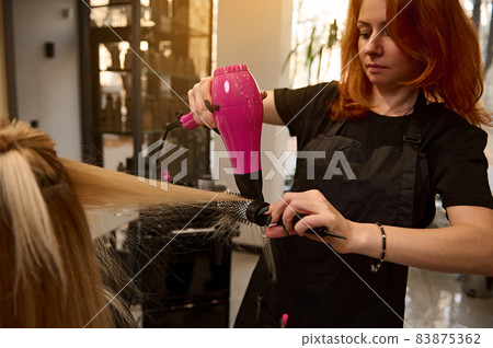 A beautiful red-haired hairdresser using a round comb and a hair dryer straightens blonde hair to a client of a beauty salon, performing her a stylish hairstyle A beautiful red-haired hairdresser using a round comb and a hair dryer straightens blonde hair to a client of a beauty salon, performing her a stylish hairstyle 83875362