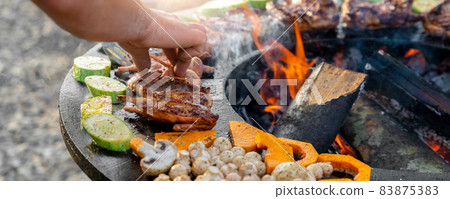 Close-up detail view of chef flipping by tongs tasty crispy marinated quails and vegetables grilled at round steel iron firepit hearth table surface brazier with burning firewoods. Barbecue yard home Close-up detail view of chef flipping by tongs tasty crispy marinated quails and vegetables grilled at round steel iron firepit hearth table surface brazier with burning firewoods. Barbecue yard home 83875383
