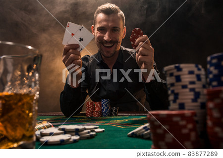 Handsome poker player with two aces in his hands and chips sitting at poker table in a dark room full of cigarette smoke. Handsome poker player with two aces in his hands and chips sitting at poker table in a dark room full of cigarette smoke. 83877289