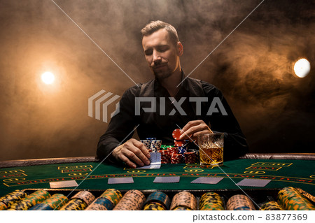 Man is playing poker with a cigar and a whiskey. A man winning all the chips on the table with thick cigarette smoke. Man is playing poker with a cigar and a whiskey. A man winning all the chips on the table with thick cigarette smoke. 83877369