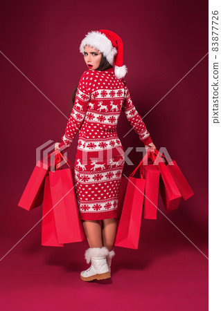 Young pretty woman or girl with long beautiful hair in Santa hat and New Year's dress holding red package on red studio background. Full-length photo Young pretty woman or girl with long beautiful hair in Santa hat and New Year's dress holding red package on red studio background. Full-length photo 83877726