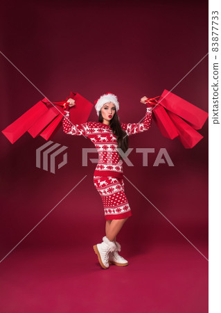 Young pretty woman or girl with long beautiful hair in Santa hat and New Year's dress holding red package on red studio background. Full-length photo Young pretty woman or girl with long beautiful hair in Santa hat and New Year's dress holding red package on red studio background. Full-length photo 83877733