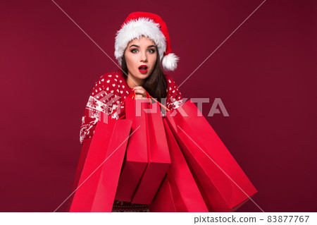 Young pretty woman or girl with long beautiful hair in Santa hat and New Year's dress holding red package on red studio background 83877767