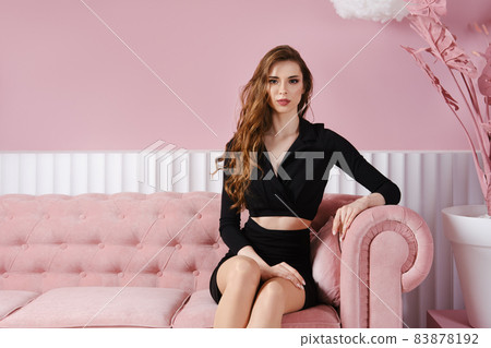 Portrait of beautiful young woman in black dress sits on sofa in pink room Portrait of beautiful young woman in black dress sits on sofa in pink room 83878192
