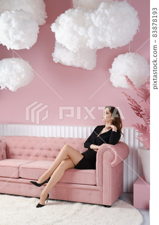 Beautiful young woman in black dress sits on sofa in pink room Beautiful young woman in black dress sits on sofa in pink room 83878193