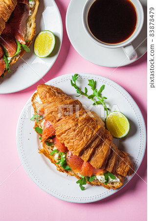 Croissant sandwich with cream cheese, salmon and arugula on white plate, pink background. Healthy breakfast concept. Croissant sandwich with cream cheese, salmon and arugula on white plate, pink background. Healthy breakfast concept. 83878479