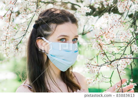 A girl in a medical face mask to avoid the spread coronavirus in the park between flowering trees. A girl in a medical face mask to avoid the spread coronavirus in the park between flowering trees. 83883613