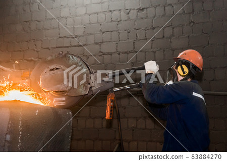 A male worker in a protective helmet, respirator, overalls manages heavy grinding equipment for cast iron concrete tubing with flying sparks in the workshop of an industrial plant 83884270