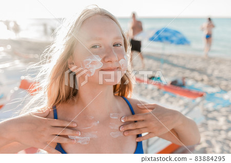 Portrait of a young positive teenage girl blonde of European appearance with sunscreen on her face and body against the background of a sea beach 83884295