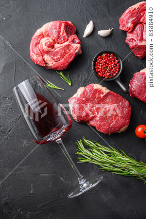 Red wine glass near raw beef steak cuts, with herbs, seasoning on black table, top view. Red wine glass near raw beef steak cuts, with herbs, seasoning on black table, top view. 83884910