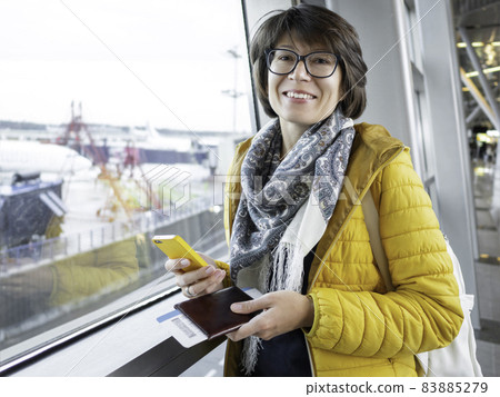 Smiling woman with passport and boarding pass is texting on her smartphone. Tourist is waiting for boarding at window at airport. 83885279