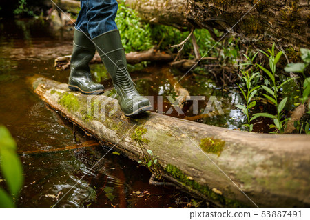 A man in rubber boots walks along a fallen tree 83887491