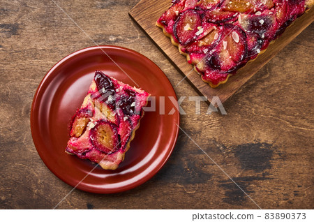Piece of plum tart on ceramic plate, next to pie on wooden board Piece of plum tart on ceramic plate, next to pie on wooden board 83890373