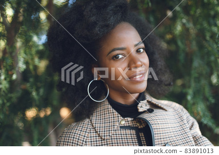 Beautiful African American young woman with afro and large hoop earrings in a stylish coat in a spring or autumn park, smiling. Selective focus, copy space Beautiful African American young woman with afro and large hoop earrings in a stylish coat in a spring or autumn park, smiling. Selective focus, copy space 83891013