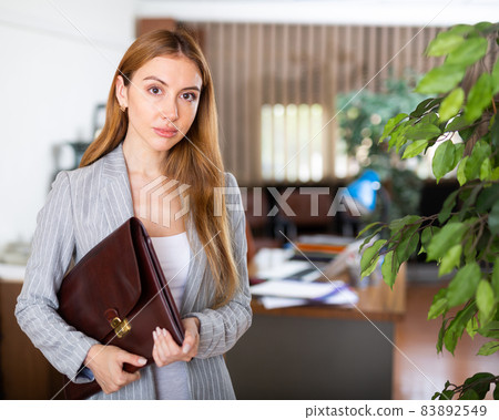 Young confident businesswoman is standing with a folder of documents in a office Young confident businesswoman is standing with a folder of documents in a office 83892549