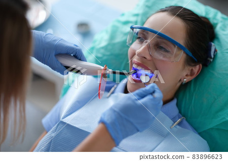 Young woman dentist in protective gloves checking dental seal, fixing photopolymer lamp during stomatology procedure Young woman dentist in protective gloves checking dental seal, fixing photopolymer lamp during stomatology procedure 83896623