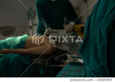 Scrub nurse hand holding scalpel for operation with surgeon performing in background. 83899897