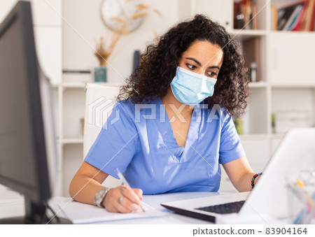 Female therapist in protective mask working at table with laptop in office 83904164