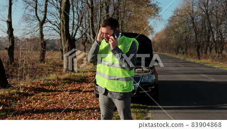 Unhappy businessman in a safety vest standing speak on phone near the broken car opened the hood road help repair. Unhappy businessman in a safety vest standing speak on phone near the broken car opened the hood road help repair. 83904868