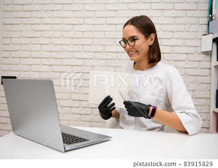 Beautiful young female doctor, general practitioner consults online while sitting at the table in hospital and showing a notebook to a patient at laptop web camera. Online medical consultation concept Beautiful young female doctor, general practitioner consults online while sitting at the table in hospital and showing a notebook to a patient at laptop web camera. Online medical consultation concept 83915829