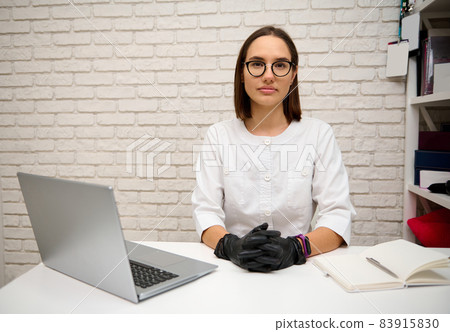 Beautiful young female doctor, general practitioner consults online while sitting at the table in hospital and confidenty looking at camera. Online medical consultation concept 83915830