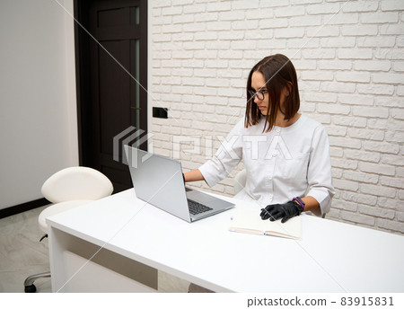 European young beautiful professional general practitioner woman, female doctor working in medical cabinet texting and browsing online on laptop sitting at desk in hospital at workplace. 83915831