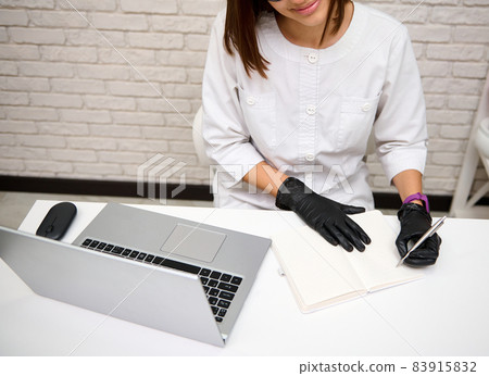 High angle view of a woman doctor, general practitioner in white medical uniform sitting at desk in hospital writing and consulting patient online on laptop, smiles confidently looking at camera High angle view of a woman doctor, general practitioner in white medical uniform sitting at desk in hospital writing and consulting patient online on laptop, smiles confidently looking at camera 83915832