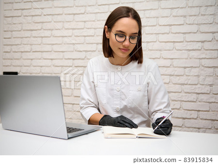 Beautiful woman general practitioner, physician, doctor in medical gown working on a laptop, writing on notepad, while having a video conferencing with a team or patient giving online treatment advice Beautiful woman general practitioner, physician, doctor in medical gown working on a laptop, writing on notepad, while having a video conferencing with a team or patient giving online treatment advice 83915834