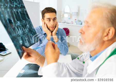 Close-up of mature adult male physician consult scared young man patient giving bad news explaining results of MRI image. 83916598