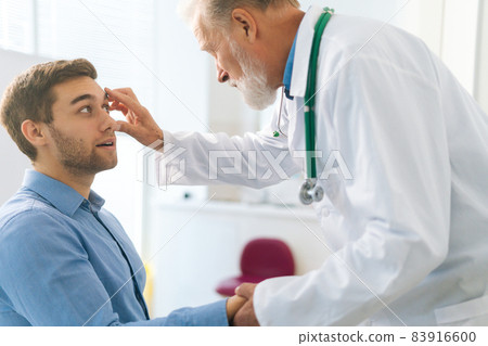 Close-up of mature adult male ophthalmologist examining eyes of male patient during checkup visit in clinic office. Close-up of mature adult male ophthalmologist examining eyes of male patient during checkup visit in clinic office. 83916600
