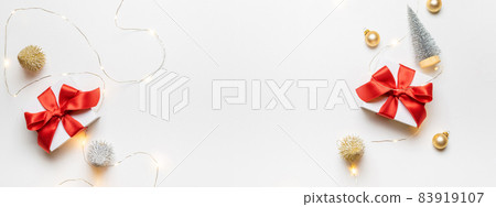 Christmas gift. White gift with red bow, gold balls and sparkling lights garland in xmas decoration on white background for greeting card. Xmas backdrop with space for text. Christmas gift. White gift with red bow, gold balls and sparkling lights garland in xmas decoration on white background for greeting card. Xmas backdrop with space for text. 83919107