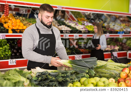 Male shop assistant in apron lays fresh leek in supermarket 83920415