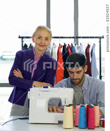 Senior fashion designer closely control the new designer in his work that require electric sewing machine. Man and woman working in home base cloth design business. 83924937