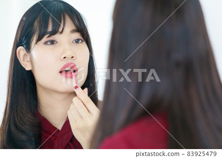 Horizontal portrait shot of attractive young adult Asian woman with long straight hairstyle in casual red clothes looking at the mirror and dyes her lips with lipstick. Woman applied makeup on face Horizontal portrait shot of attractive young adult Asian woman with long straight hairstyle in casual red clothes looking at the mirror and dyes her lips with lipstick. Woman applied makeup on face 83925479