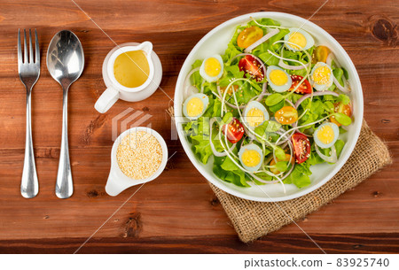 Big white bowl of lettuce, sprout salad with boil egg, onion and tomato mix with Japanese sesame oil dressing on wooden table together with small olive oil jar and sesame cup and utensil beside Big white bowl of lettuce, sprout salad with boil egg, onion and tomato mix with Japanese sesame oil dressing on wooden table together with small olive oil jar and sesame cup and utensil beside 83925740