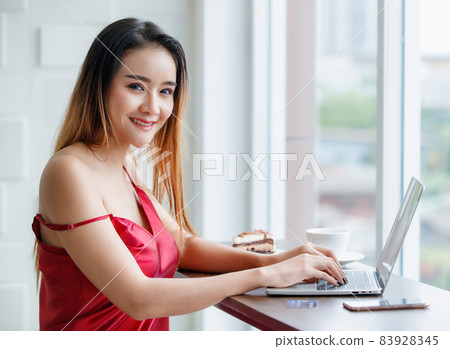 Beautiful young cut ASian sexy girl on red seductive dress lovely smile to other as she enjoying work on laptop in bright morning with breakfast of piece of cake and cup of coffee 83928345