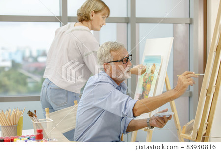 Caucasian old senior elderly male and female artist painter drawing painting flower bouquet picture with watercolor in palette and paintbrush on canvas paper on easel stand together in hobby workshop Caucasian old senior elderly male and female artist painter drawing painting flower bouquet picture with watercolor in palette and paintbrush on canvas paper on easel stand together in hobby workshop 83930816