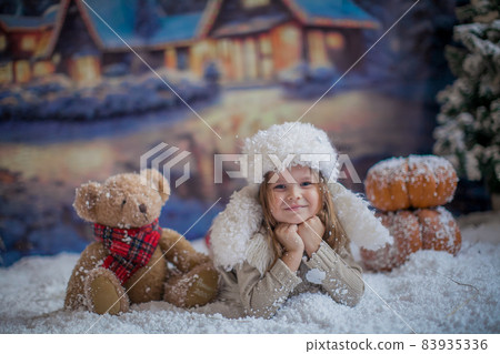 A girl in a warm winter white hat lies on her stomach next to a toy bear cub on the background of a winter village A girl in a warm winter white hat lies on her stomach next to a toy bear cub on the background of a winter village 83935336