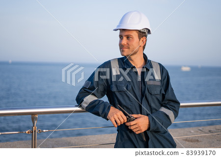 Confident smiling young bearded male worker in protective clothes and hardhat with walkie talkie in hands leaning against metal railing and looking away while having break during work in sea port 83939170