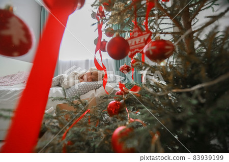 Photo of a boy in pajamas on a bed next to a Christmas tree decorated with red toys Photo of a boy in pajamas on a bed next to a Christmas tree decorated with red toys 83939199