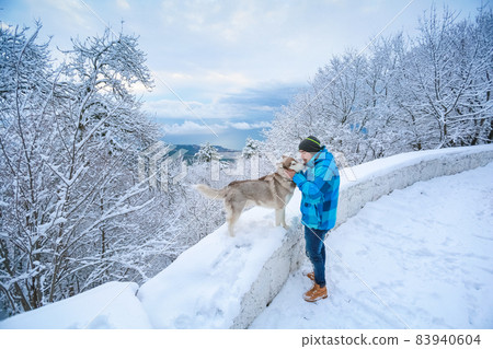 A man in warm clothes stands with a dog along a snowy mountain road A man in warm clothes stands with a dog along a snowy mountain road 83940604