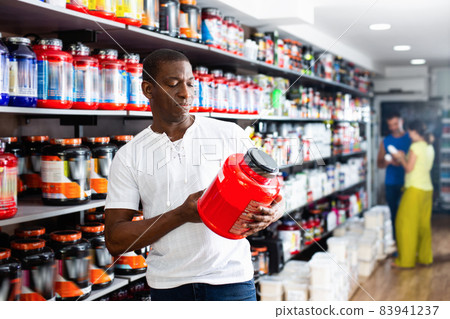 Focused muscular African man choosing sports nutrition products in shop, reading content label Focused muscular African man choosing sports nutrition products in shop, reading content label 83941237
