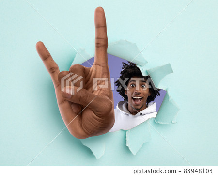 Excited black teenager showing huge rock gesture through hole in light blue ripped paper 83948103