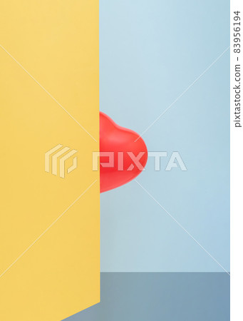 Red heart-shaped balloon hides behind a yellow wall in front of a blue background Red heart-shaped balloon hides behind a yellow wall in front of a blue background 83956194